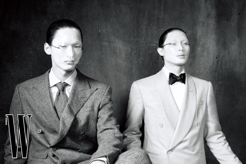 Two East Asian male models in tailored double-breasted suits and translucent face shields in a black and white studio portrait.