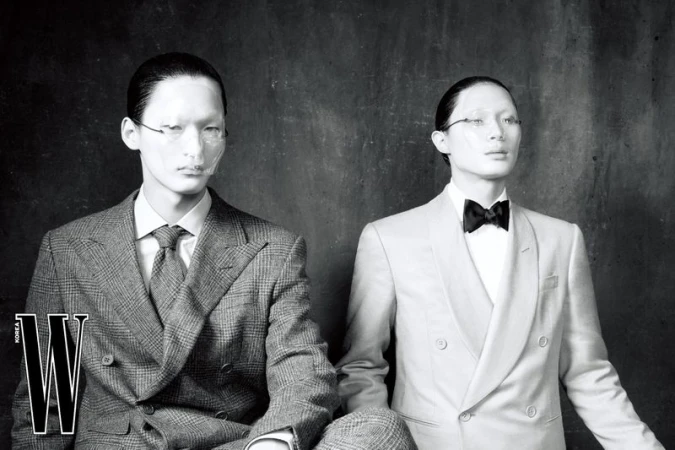 Two East Asian male models in tailored double-breasted suits and translucent face shields in a black and white studio portrait.