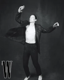 Model Jinyeong Pak posing shirtless in an open dark suit jacket and trousers for W Korea against a dark textured backdrop.