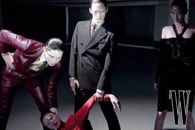 Three models in dark tailored suits and a black dress posing dramatically under studio lighting for W Korea editorial photography.