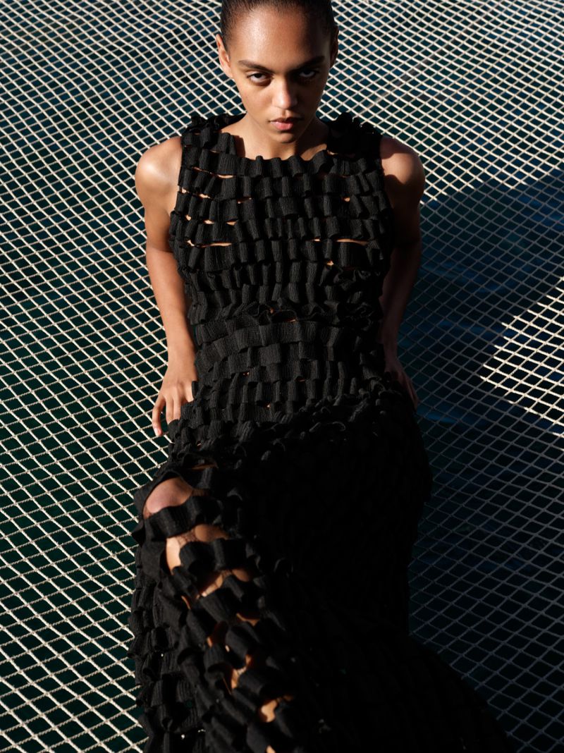Model Bebe Parnell reclining on an open mesh net wearing a textured black woven dress in direct sunlight.