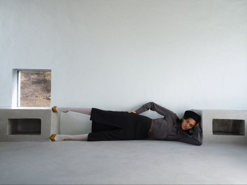 Model lying horizontally along a white concrete ledge wearing a dark shirt, black trousers, white tights, and yellow heels