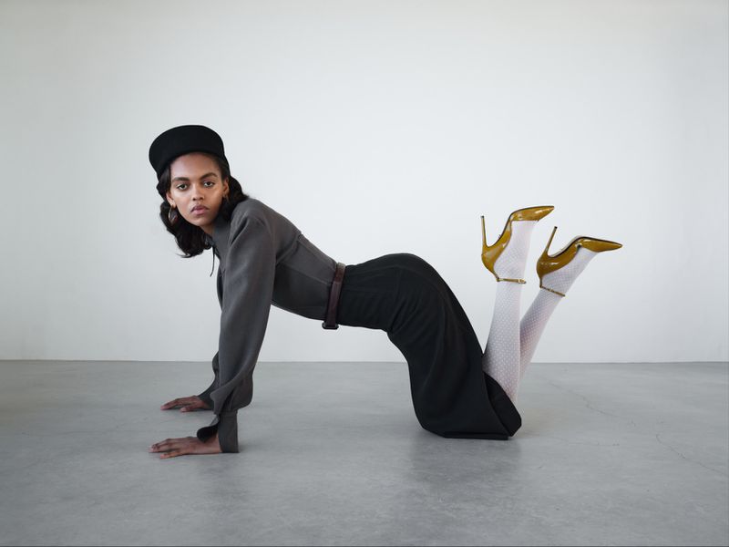 Model Bebe Parnell poses in a grey blouse, black skirt, white tights, and yellow patent heels on a grey studio floor.