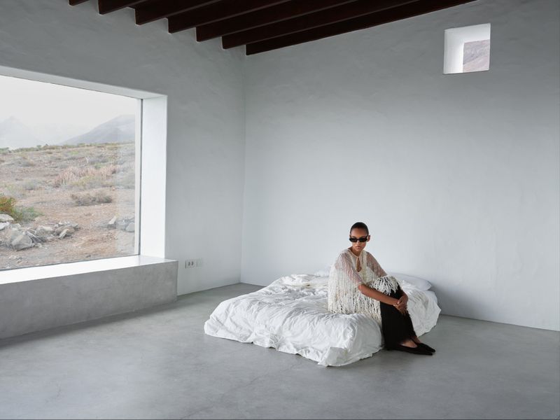 A model wearing a white fringed top and sunglasses sits on a low white bed inside a minimalist minimalist concrete room with a large window.
