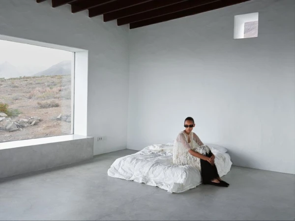 A model wearing a white fringed top and sunglasses sits on a low white bed inside a minimalist minimalist concrete room with a large window.