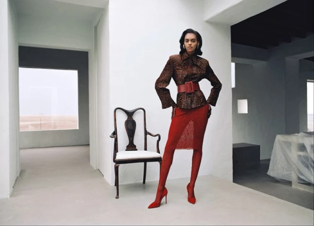 Model Bebe Parnell in a brown jacket, red belt, red skirt, and red heels posing by an antique armchair in a bright white space.