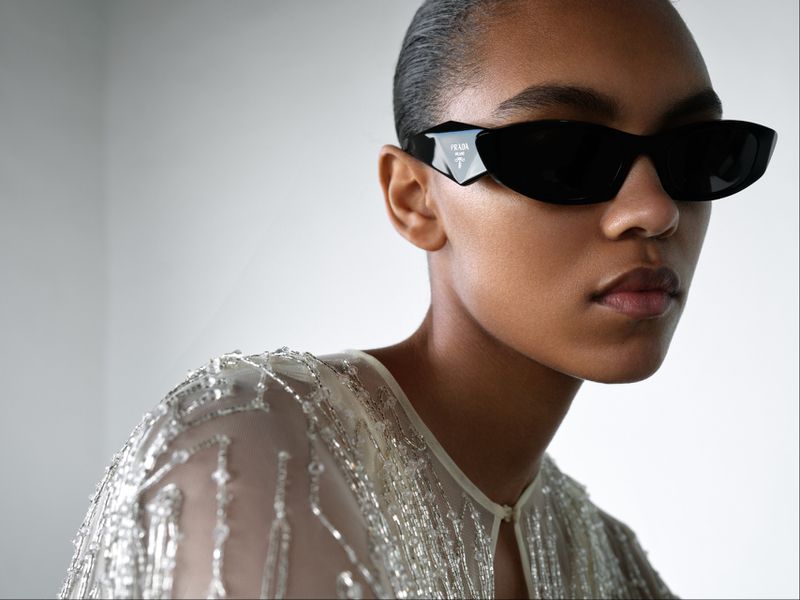 Close-up of a person wearing black Prada sunglasses and a beaded top against a light background in a studio shot. Fashion editorial.