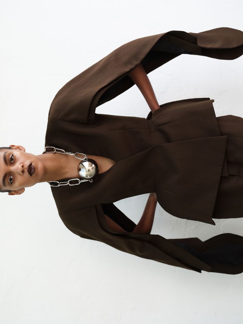 Model wearing a dark brown sculptural jacket and oversized silver chain necklace against a white background