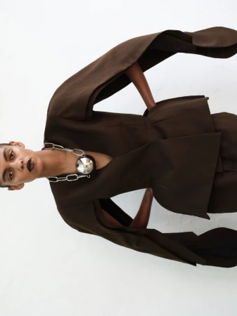 Model wearing a dark brown sculptural jacket and oversized silver chain necklace against a white background