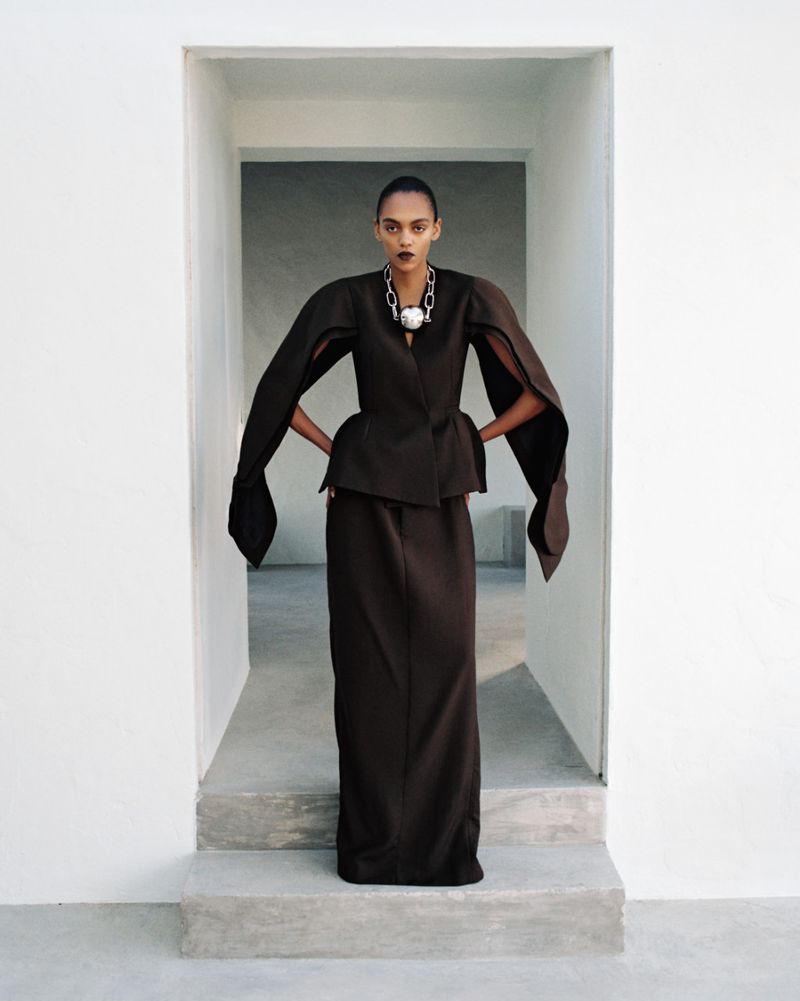 Model Bebe Parnell posing in a sculptural dark brown outfit with dramatic slit sleeves within a white architectural doorway.