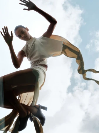 Low-angle shot of model Bebe Parnell wearing a cream draped top and flowing ribbons against a cloudy sky.