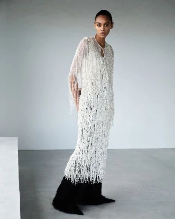 Model Bebe Parnell posing in a white beaded fringe dress and black pointed boots against a neutral grey studio background.