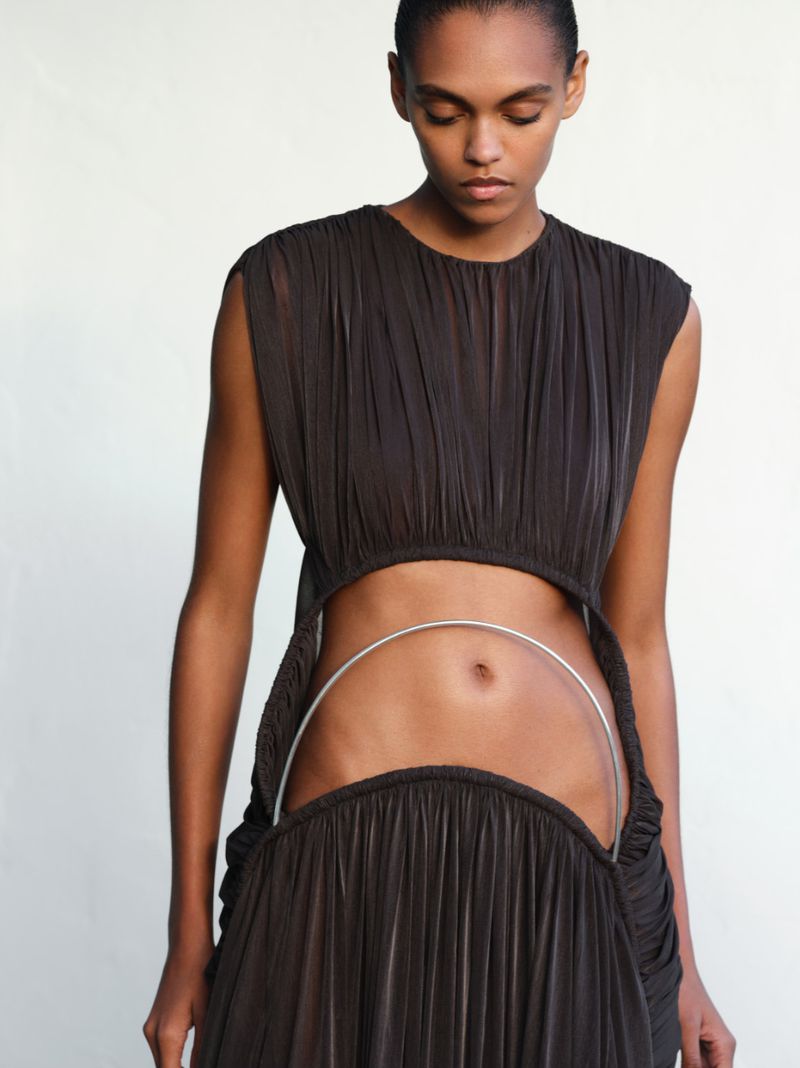Model Bebe Parnell wearing a dark brown pleated cutout dress with a curved metal midriff detail against a white background.
