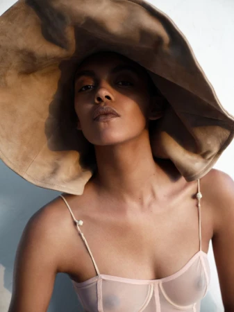 Model Bebe Parnell wearing an oversized floppy camel hat and a sheer pale pink corset top against a neutral backdrop.