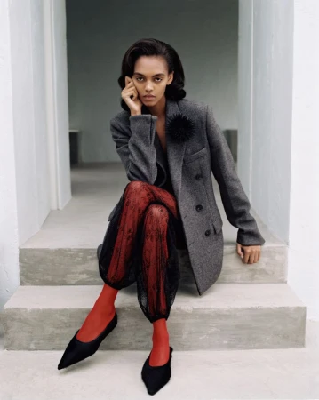 Model Bebe Parnell seated on concrete steps wearing a grey wool blazer, black sheer lace pants, red tights, and pointed black flats.