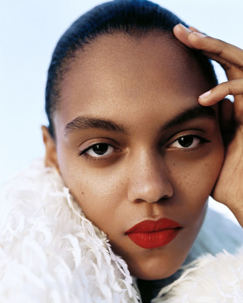 Close-up portrait of model Bebe Parnell with bold red lipstick and a white textured feathered collar against a pale blue sky.