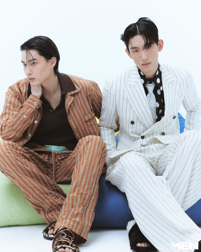 Two male models seated in a striped brown suit and a white pinstripe double-breasted suit against a white studio background.