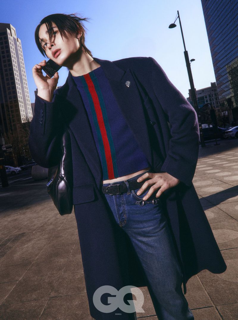 Model in a dark navy coat and striped crop top holding a phone to his ear in a city plaza.