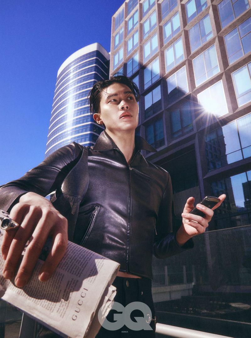 Urban fashion shot of a man in a leather jacket holding a phone and newspaper in front of modern buildings under a bright blue sky.