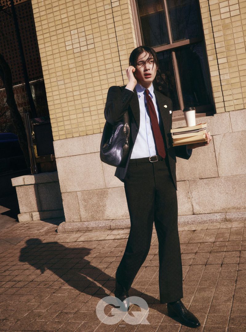 Model Jinyeong Pak in a tailored black suit and red tie talking on a mobile phone while carrying books on a sunny sidewalk.