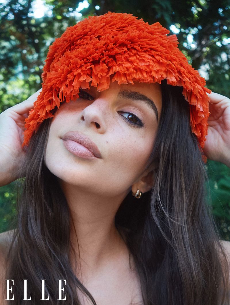 Portrait of a woman with brown hair wearing orange floral headwear, gazing at the camera with a neutral expression in an outdoor setting.