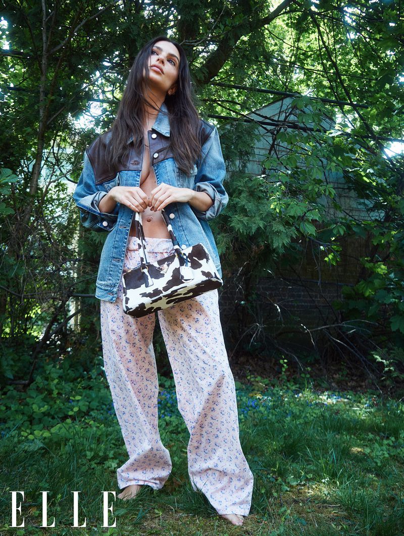 Woman posing outdoors in a denim jacket, patterned pants, and cow-print bag, surrounded by green foliage, captured at eye-level.