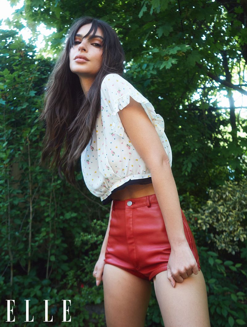 Emily Ratajkowski wearing a white patterned top and red shorts outdoors against a lush green leafy background for Elle UK.