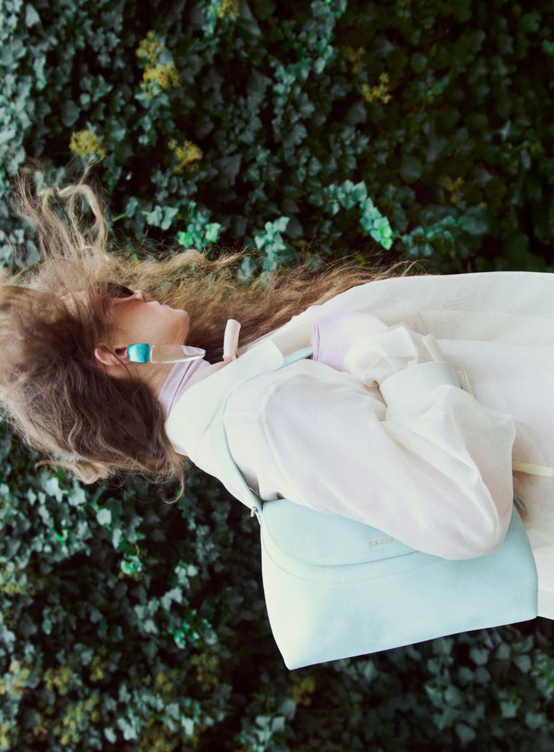 Woman in an off-white jacket with wavy hair carrying a light blue shoulder bag against a background of dense green foliage.