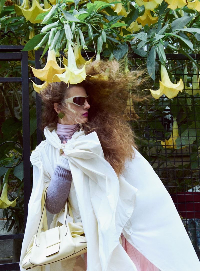 Model in sunglasses wearing a white cape and holding a pale yellow handbag beneath hanging yellow flowers for D Repubblica by Joan Braun.