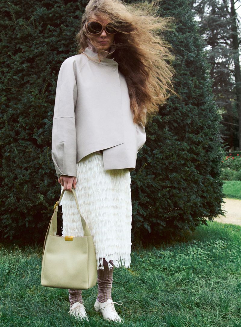 Woman with windblown hair wearing a pale grey jacket, white fringe skirt, and oversized sunglasses holding a tote bag on a lawn.