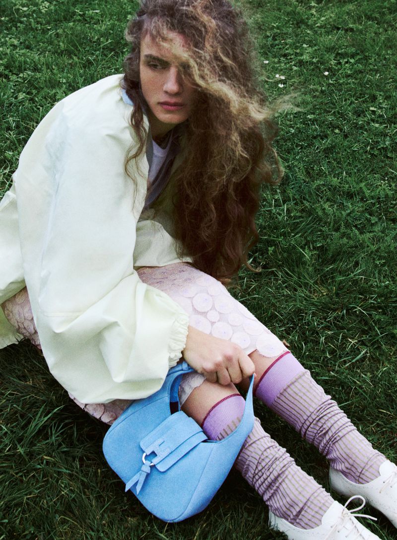 Model with long wavy hair seated on green grass holding a light blue handbag, wearing an off-white jacket and lilac socks.