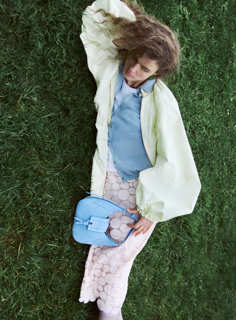 Overhead shot of a woman lying on green grass in a yellow jacket, blue top, patterned pants, and holding a blue handbag.