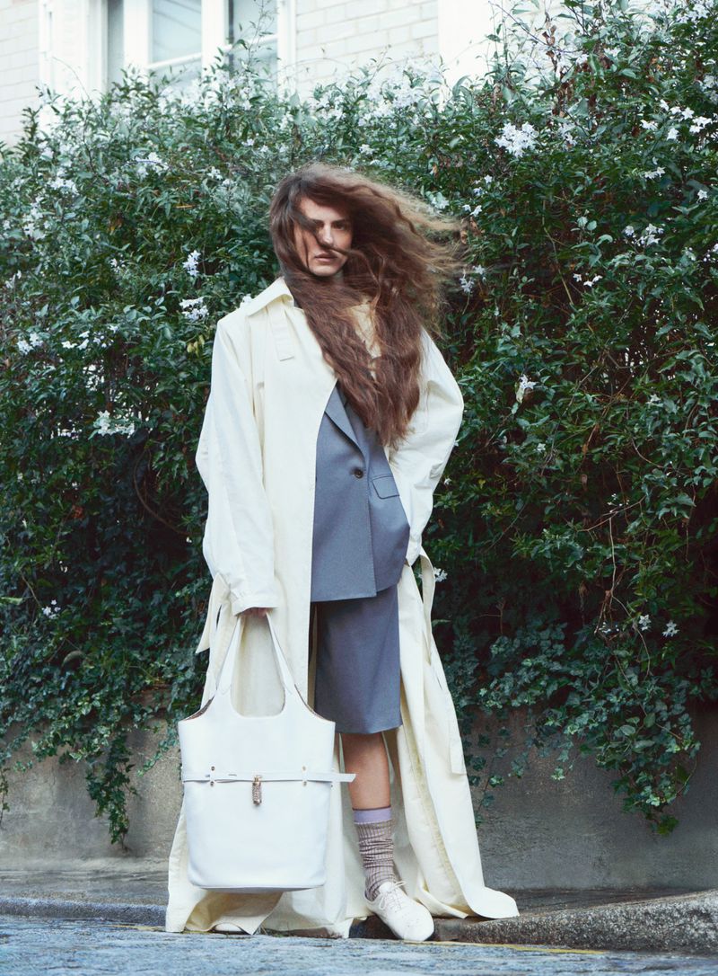 Model with long windblown hair wearing a white trench coat and grey blazer outdoors against green foliage
