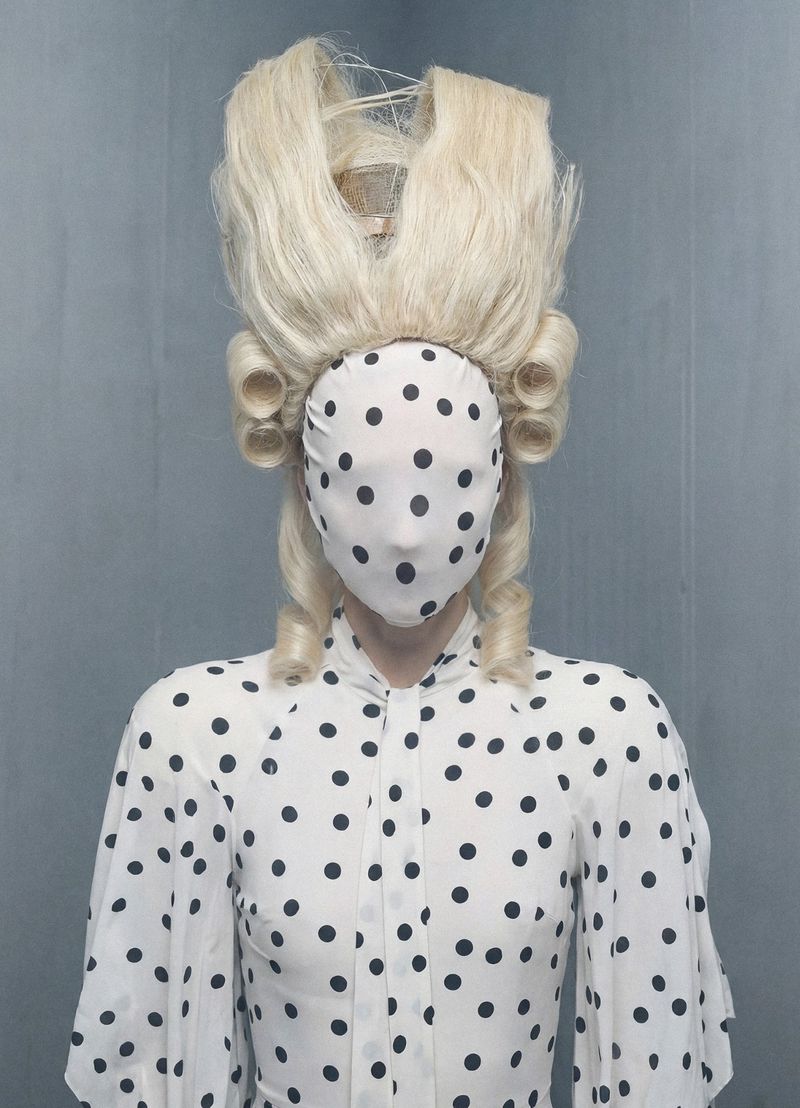 Figure wearing a polka dot face covering and matching blouse paired with a high sculptural blonde wig against a grey backdrop.
