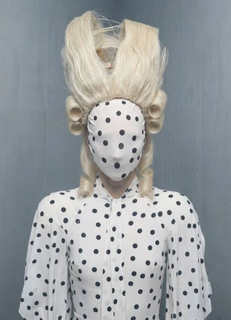 Figure wearing a polka dot face covering and matching blouse paired with a high sculptural blonde wig against a grey backdrop.