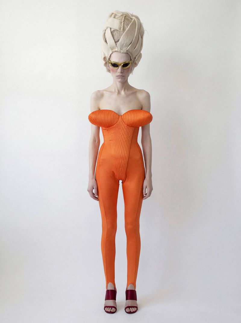 Model wearing a strapless orange jumpsuit, high sculptural blonde wig, and sunglasses against a seamless white studio backdrop.