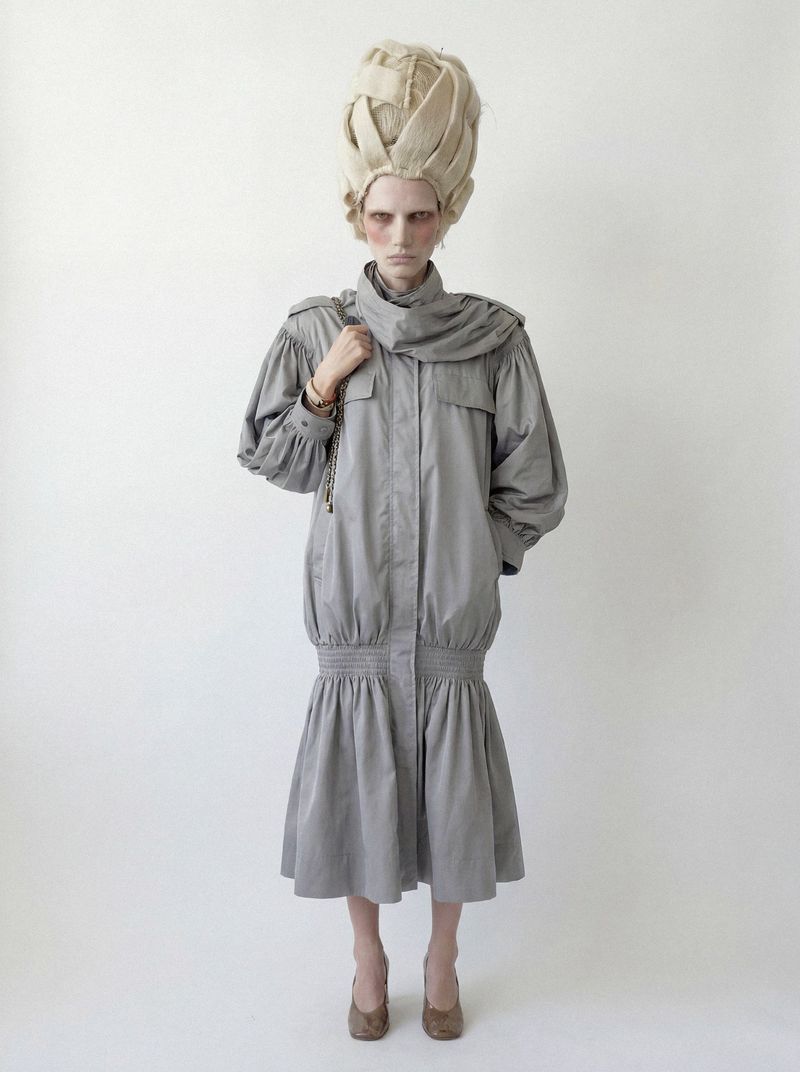 Model wearing a grey drop-waist gathered coat dress and a tall sculpted blonde headpiece against a plain white backdrop.