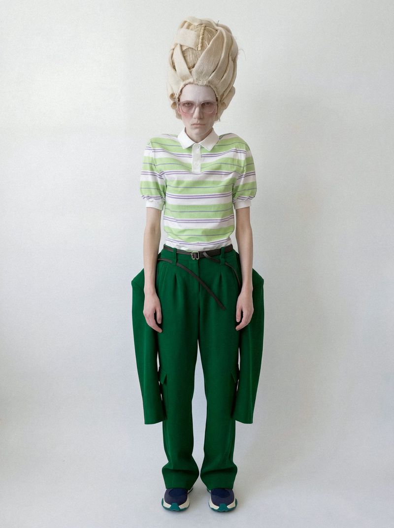 A fashion model wearing a sculptural blonde wig, green striped polo shirt, and green trousers against a white seamless studio backdrop.