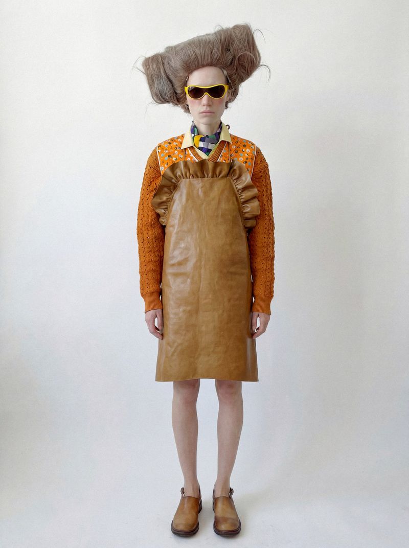 Fashion model in an oversized sculpted wig, yellow sunglasses, and a brown leather apron dress against a white studio backdrop.