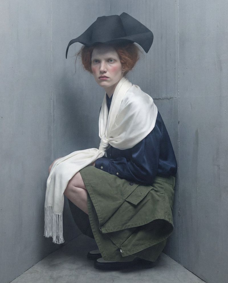 Model wearing a black sculptural hat, white scarf, and olive skirt crouched in a corner for Vogue Czechoslovakia by Polina and Vladimir Mordvinovs