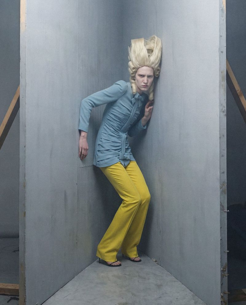 Model in blue jacket and yellow trousers wearing a towering blonde wig in a grey corner studio set