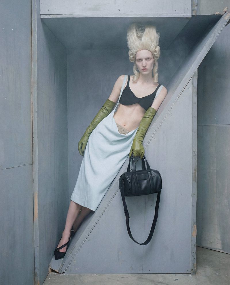 Fashion model with towering blonde hair in a black top, pale blue drape skirt, and olive leather gloves leaning in a grey alcove.