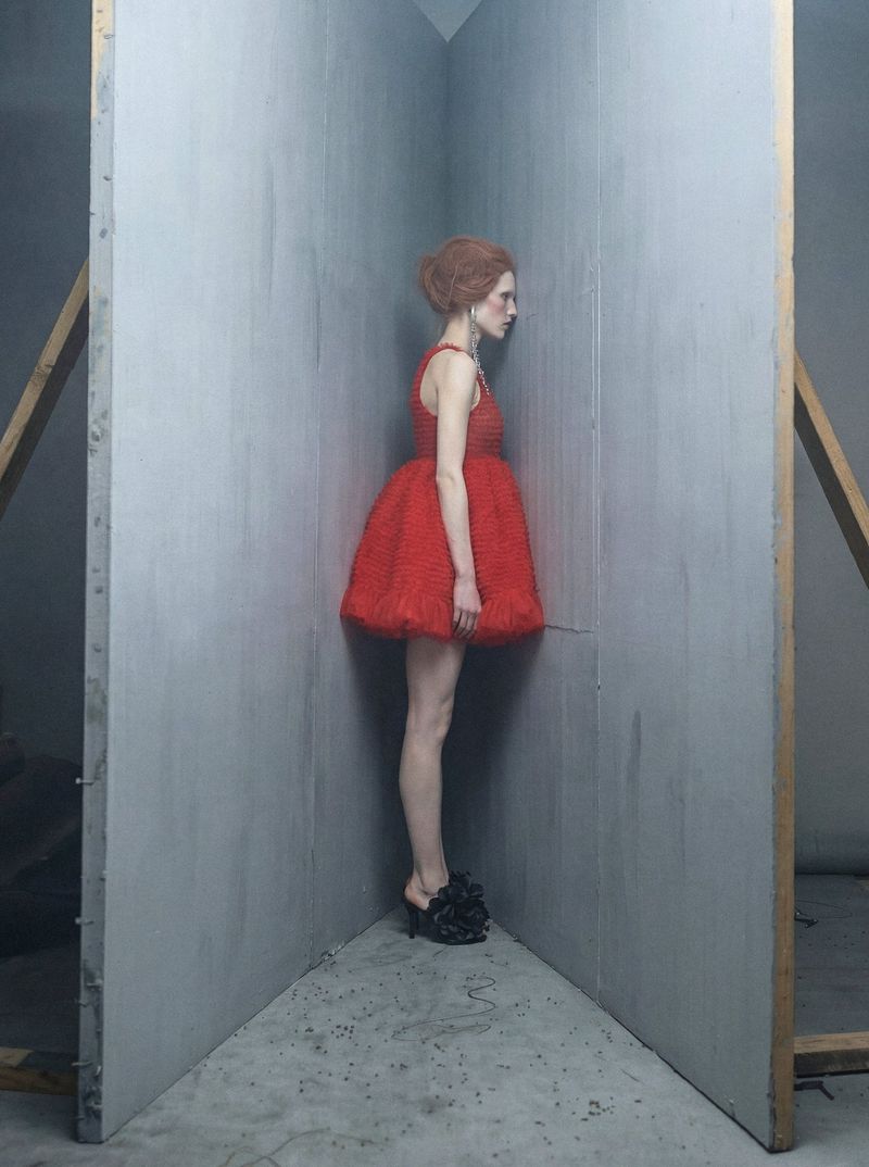 A slender woman with auburn hair poses in a red dress and platform heels inside a geometric corner space with grey walls and wooden beams.