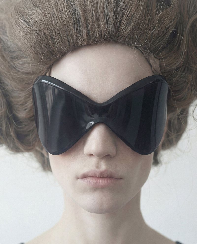 Close-up portrait of a model wearing oversized black sunglasses and dramatic teased brown hair against a white background