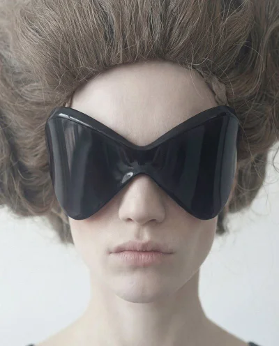 Close-up portrait of a model wearing oversized black sunglasses and dramatic teased brown hair against a white background