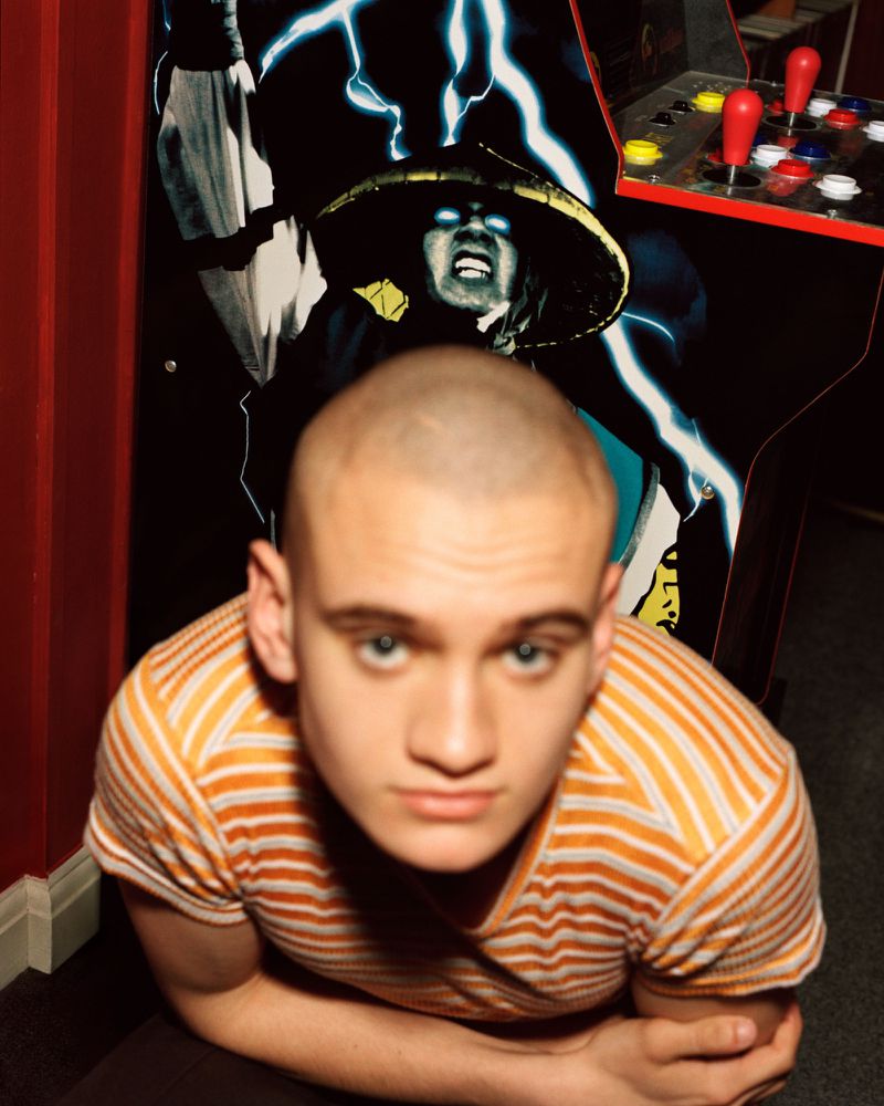 Close-up portrait of a young man with a buzz cut in an orange striped shirt standing in front of an arcade cabinet.