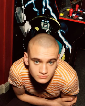 Close-up portrait of a young man with a buzz cut in an orange striped shirt standing in front of an arcade cabinet.