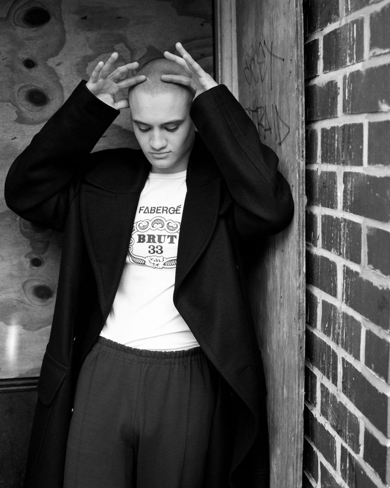 Monochrome portrait of young man in black coat and graphic t-shirt leaning against a brick wall with hands on head.