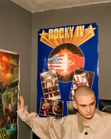 A young man with a shaved head in a textured collared jacket poses against a wall with a Rocky IV movie poster.