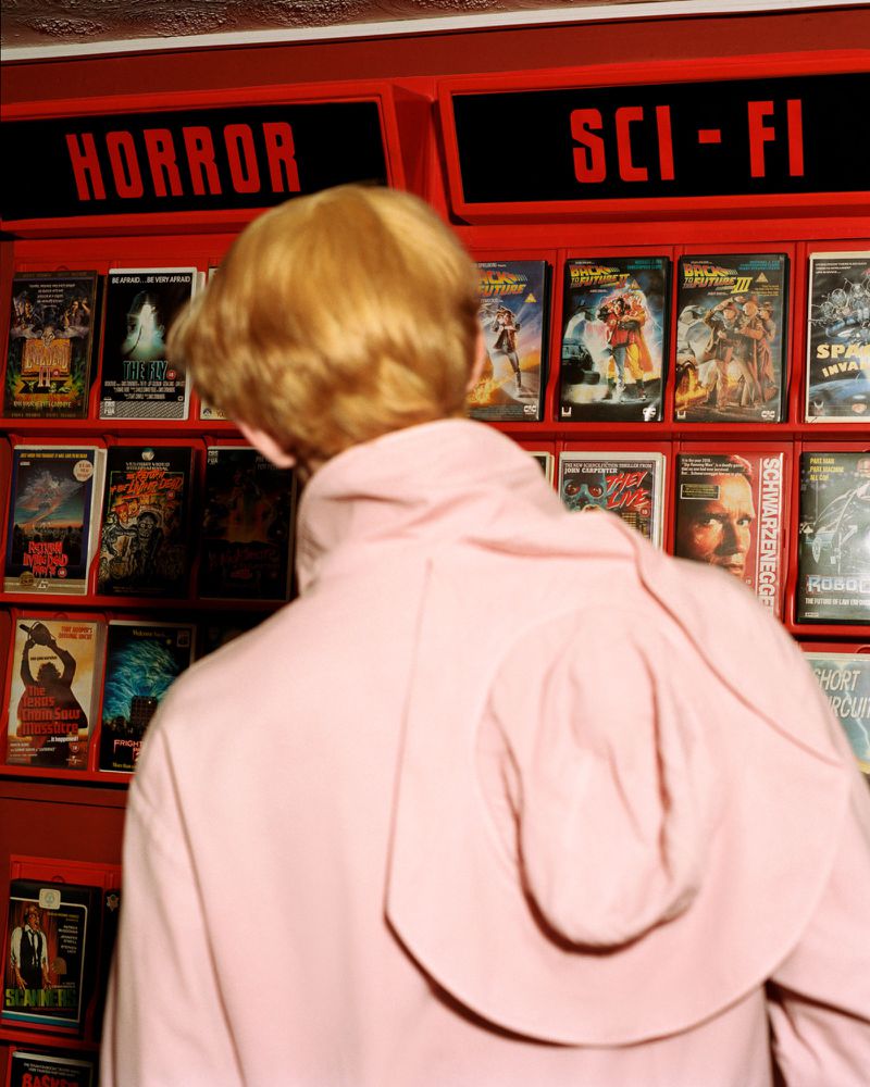 A person with blond hair seen from behind in a pale pink sweatshirt standing in front of shelves filled with VHS tapes.
